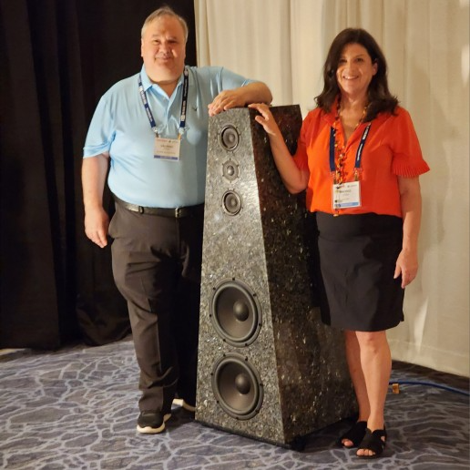 Two people standing beside a tall speaker with multiple drivers.