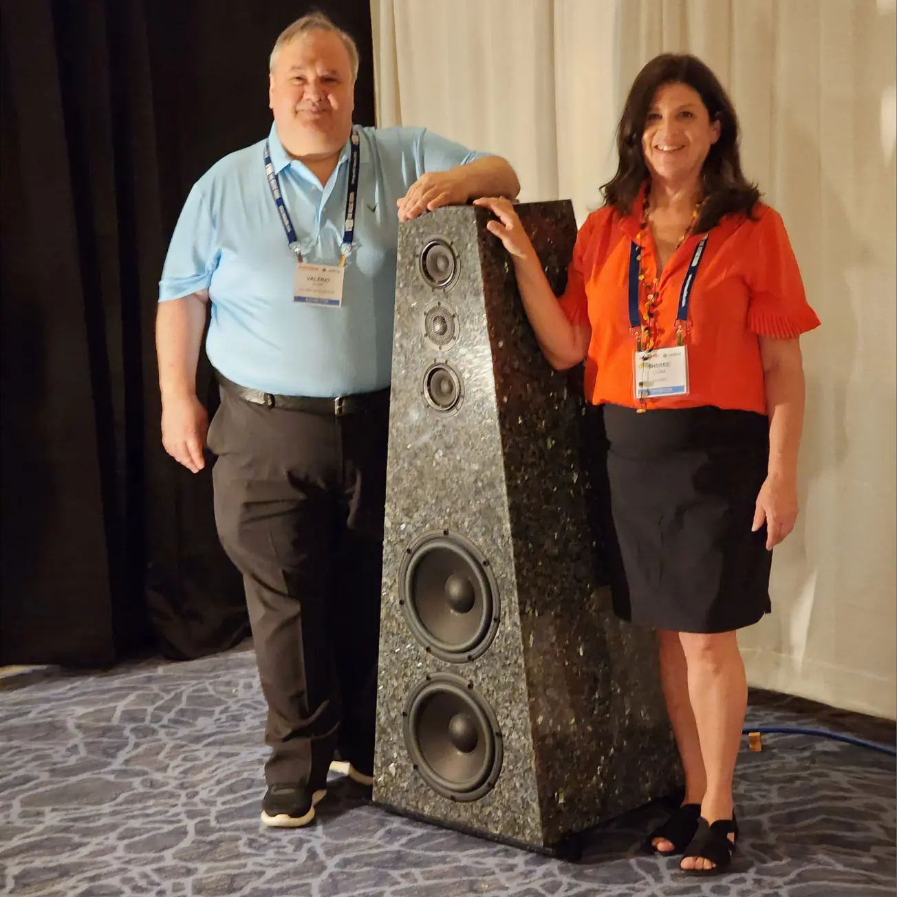 Two people posing with a large speaker in a room.