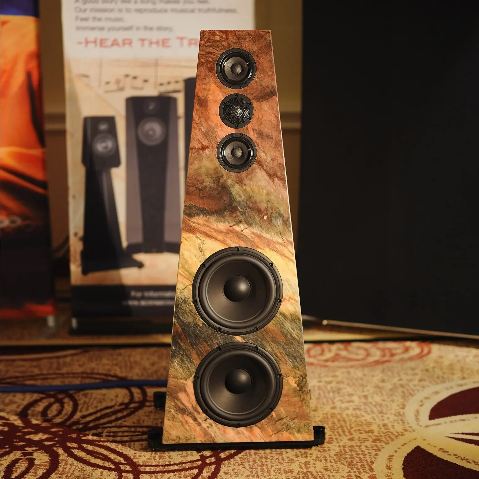 Tall wooden speaker with multiple drivers on a patterned carpet.