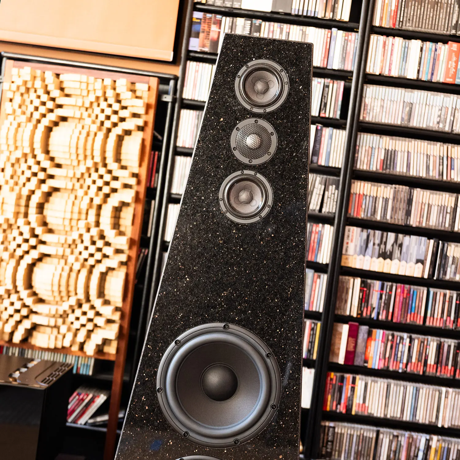 Tall black speaker with multiple drivers in a room filled with CDs and patterned decor.