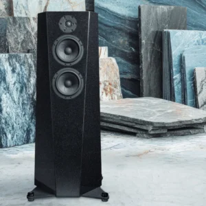 Black floor-standing speaker with dual drivers in a stone workshop.